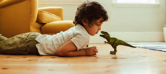 Niedlicher kleiner Junge, der sein Dinosaurierspielzeug zu Hause betrachtet. Niedlicher kleiner Junge, der mit einem T-Rex-Dinosaurierspielzeug spielt, während er auf dem Boden in seinem Spielbereich liegt. - JLPSF11246