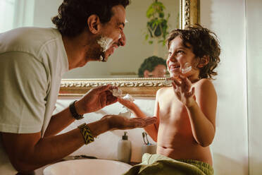 Father and son having fun with shaving foam. Happy father and son smiling at each other with shaving cream on their faces. Loving young father bonding with his son in the bathroom at home. - JLPSF11247