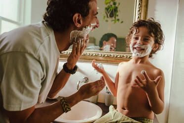 Happy father and son having fun with shaving cream in the bathroom. Father and son smiling cheerfully with shaving foam on their faces. Loving father bonding with his young son at home. - JLPSF11249