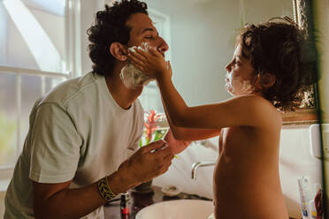 Sweet young boy applying shaving cream on his father's face. Adorable young boy helping his father shave his facial hair in the bathroom. Loving father and son bonding at home. - JLPSF11250