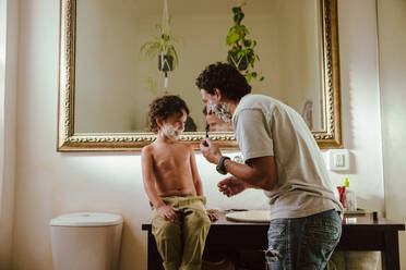 Young father and son having fun with shaving cream in the bathroom. Happy father and son smiling at each other with shaving foam on their faces. Loving father bonding with his young son at home. - JLPSF11254