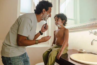 Cheerful father showing his son how to shave at home with shaving cream. Happy father teaching his son his shaving skills in the bathroom. Father and son spending some quality time together. - JLPSF11255