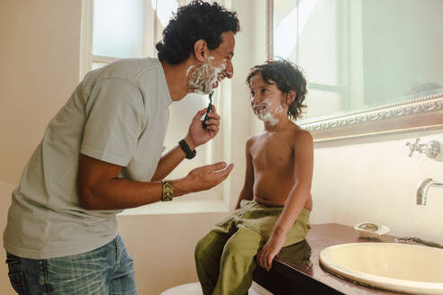 Ein fröhlicher Vater, der seinem Sohn zeigt, wie man sich zu Hause mit Rasierschaum rasiert. Ein glücklicher Vater, der seinem Sohn im Badezimmer seine Rasierkünste beibringt. Vater und Sohn verbringen eine schöne Zeit miteinander. - JLPSF11255