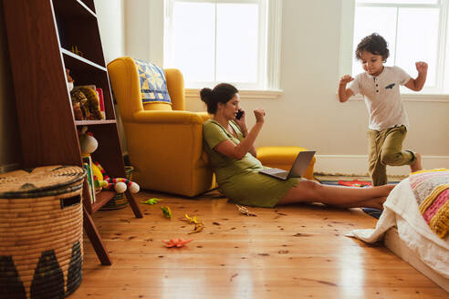 Self-employed mother speaking on the phone while sitting in her son's play area. Working mother making plans with her clients over a phone call. Young single mother working from home. - JLPSF11259