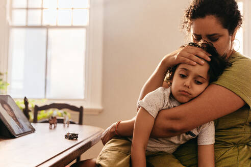 Compassionate mother caring for her sick son at home. Loving mother embracing her young son during an online consultation with their family doctor. Mother and son video calling their paediatrician. - JLPSF11263