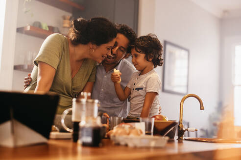 Verspielte Familie, die gemeinsam in der Küche eine Banane isst. Glücklicher Junge, der seine Eltern mit einer Banane füttert, während er auf dem Küchentisch sitzt. Mutter und Vater verbringen viel Zeit mit ihrem Sohn. - JLPSF11274