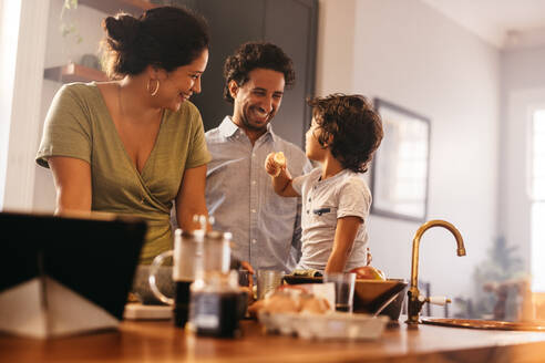 Familienzeit in der Küche: Ein glücklicher Junge isst eine Banane, während er auf dem Küchentisch sitzt. Mama und Papa lächeln glücklich, während sie mit ihrem Sohn zu Hause Zeit verbringen. - JLPSF11275