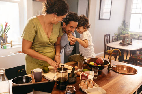 Eine dreiköpfige Familie verbringt viel Zeit in der Küche. Der verspielte Vater isst eine Scheibe Brot, während seine Frau das Frühstück zubereitet. Eine junge Familie, die morgens zusammen Spaß hat. - JLPSF11279