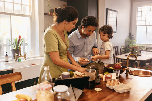 Eine Familie frühstückt gemeinsam zu Hause. Eine junge ethnische Familie bestreicht ihr Brot mit Marmelade in der Küche. Zwei liebevolle Eltern verbringen viel Zeit mit ihrem kleinen Sohn. - JLPSF11281