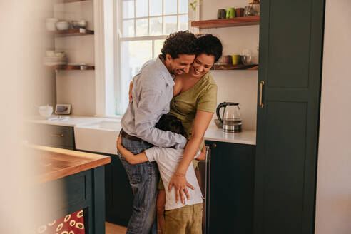 Happy ethnic family embracing each other affectionately. Sweet little boy greeting his parents with a warm hug in the kitchen. Cheerful family of three having some quality time at home. - JLPSF11283