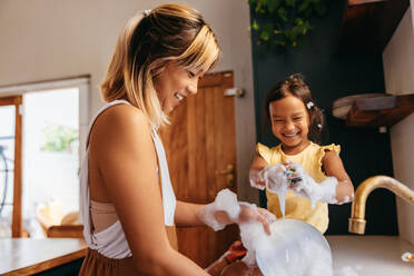 Glückliche Mutter und Tochter waschen Geschirr mit Blasen. Mutter und Tochter haben Spaß zusammen in der Küche. Liebevolle alleinerziehende Mutter verbringt Zeit mit ihrer kleinen Tochter zu Hause. - JLPSF11295