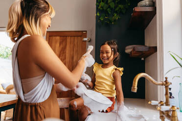 Mutter und Tochter haben Spaß mit Seifenschaum in der Küche. Glückliches kleines Mädchen lacht mit ihrer Mutter, während sie auf der Arbeitsplatte des Waschbeckens sitzt. Mutter und Tochter verbringen viel Zeit miteinander. - JLPSF11298