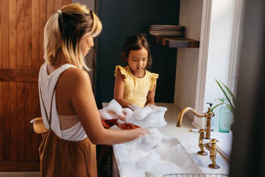 Kleines Mädchen, das lernt, wie man zu Hause abwäscht. Niedliches kleines Mädchen, das seiner Mutter beim Abwaschen hilft, während es auf der Arbeitsplatte des Waschbeckens sitzt. Mutter und Tochter verbringen viel Zeit miteinander. - JLPSF11299