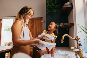 Glückliche Mutter und Tochter, die Spaß mit Seifenblasen in der Küche haben. Fröhliche Mutter, die mit ihrer Tochter beim Abwasch lacht. Liebevolle alleinerziehende Mutter, die mit ihrer Tochter zusammen ist. - JLPSF11300