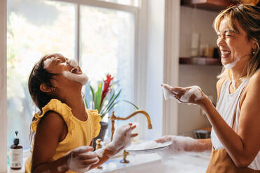 Verspielte Mutter und Tochter haben Spaß mit Seifenschaum in der Küche. Glückliches kleines Mädchen lacht fröhlich mit Seifenblasen auf ihrem Gesicht. Mutter und Tochter verbringen viel Zeit miteinander. - JLPSF11301