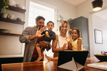 Eine bunte Familie winkt während eines Videoanrufs zu Hause. Eine glückliche ethnische Familie lächelt fröhlich, während sie ihre Lieben mit einem digitalen Tablet begrüßt. Eltern verbringen Zeit mit ihren Kindern zu Hause. - JLPSF11307