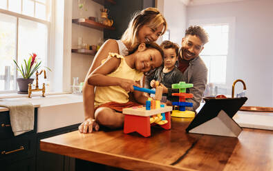 Ein Mädchen spielt mit ihren Spielsachen bei einem Videoanruf. Eine glückliche vierköpfige Familie kommuniziert mit ihren Lieben über ein digitales Tablet. Eine junge Familie lächelt fröhlich in der Küche zu Hause. - JLPSF11309