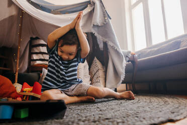 Playful little boy watching a kids programme on a digital tablet. Creative young boy putting his hands together while looking at educational content online. Ethnic boy sitting in his playing area at home. - JLPSF11315