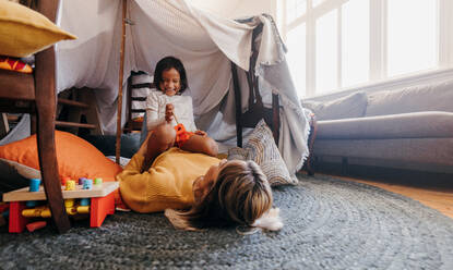 Cheerful little girl having fun with her mom during playtime. Adorable young girl laughing happily while sitting on top of her mom in her play area. Mother and daughter spending some quality time. - JLPSF11321