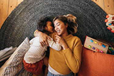 Overhead view of a mother and her daughter lying on the floor in their play area. Affectionate mother smiling happily while embracing her daughter. Mother and daughter spending quality time at home. - JLPSF11325