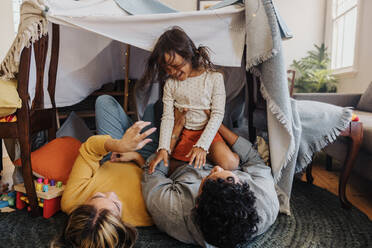 Happy little girl playing with her mom and dad at home. Adorable little girl laughing cheerfully while sitting on top of her father. Family of three spending some quality time together. - JLPSF11329