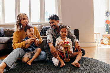 Two young parents playing with their son and daughter in the living room. Mom and dad having fun with their kids during playtime. Family of four spending some quality time together at home. - JLPSF11336