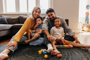 Junge Familie lächelt glücklich in ihrem Wohnzimmer zu Hause. Zwei liebevolle Eltern sitzen mit ihrem Sohn und ihrer Tochter beim Spielen. Vierköpfige Familie, die eine schöne Zeit miteinander verbringt. - JLPSF11337