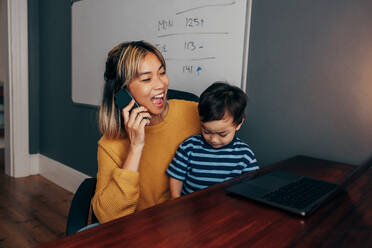 Eine fröhliche Mutter, die mit ihrem Sohn auf dem Schoß telefoniert. Eine berufstätige Mutter, die in ihrem Heimbüro mit ihren Geschäftskunden kommuniziert. Eine glückliche Mutter eines Kindes, die aus der Ferne arbeitet. - JLPSF11351