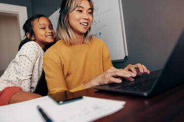 Glückliches kleines Mädchen, das seiner Mutter bei der Arbeit am Laptop zuschaut. Arbeitende Mutter, die ihrer Tochter lächelnd zeigt, wie man eine E-Mail verschickt. Fröhliche Mutter eines Kindes, die aus der Ferne arbeitet. - JLPSF11354