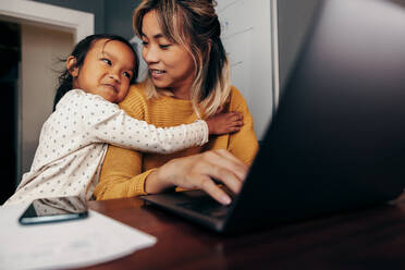 Niedliches kleines Mädchen, das seine Mutter in ihrem Büro zu Hause umarmt. Arbeitende Mutter, die ihre Tochter anlächelt, während sie eine E-Mail auf einem Laptop tippt. Selbstständige Mutter eines Kindes, die aus der Ferne arbeitet. - JLPSF11355