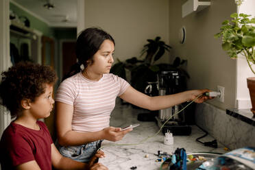 Mädchen lädt ihr Smartphone auf, während sie mit ihrem Bruder in der Nähe der Küchentheke zu Hause steht - MASF32004
