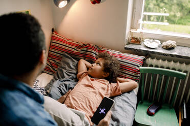 Boy looking at illuminated electric lamp while father holding smart phone on bed at home - MASF32010