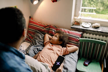 Junge schaut auf eine beleuchtete elektrische Lampe, während sein Vater sein Smartphone auf dem Bett zu Hause hält - MASF32010