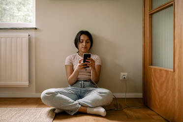 Mädchen, das ein Smartphone benutzt, während es im Schneidersitz zu Hause an der Wand sitzt - MASF32013