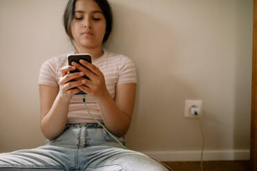 Mädchen, das sein Mobiltelefon auflädt und benutzt, während es zu Hause an der Wand sitzt - MASF32014