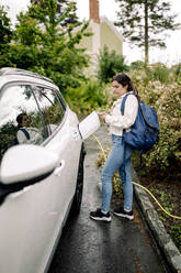 Seitenansicht einer Frau, die beim Aufladen eines Elektroautos ihr Smartphone benutzt - MASF32022