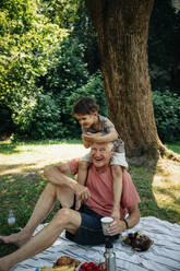 Grandfather carrying grandson on shoulder while sitting on picnic blanket at park - MASF32053