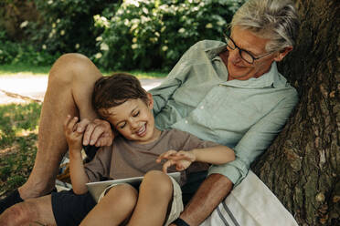 Junge benutzt digitales Tablet, während er mit seinem Großvater im Park sitzt - MASF32060