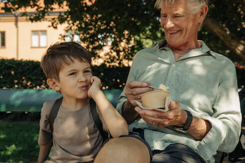 Boy leaning on elbow while sitting with grandfather holding ice cream cup at bench - MASF32073