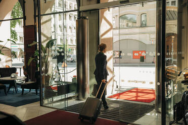 Rear view of businesswoman with suitcase leaving hotel - MASF32087