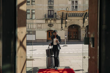 Junge Geschäftsfrau, die ein Smartphone benutzt, während sie mit einem Koffer vor einem Hotel in der Sonne steht - MASF32091