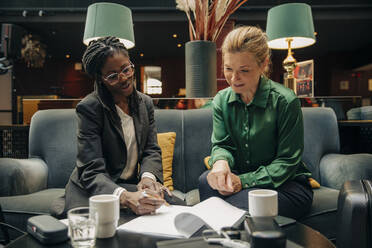Young businesswoman talking with female colleague while signing contract in hotel lounge - MASF32100