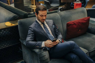 Junger Geschäftsmann, der auf dem Sofa in einer Hotellounge sitzt und über sein Smartphone SMS schreibt - MASF32107