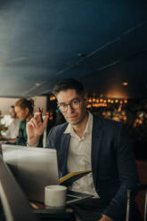 Portrait of businessman wearing eyeglasses sitting with laptop in hotel - MASF32118