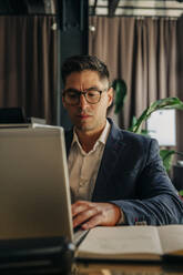 Mittlerer erwachsener Geschäftsmann mit Brille, der einen Laptop benutzt, während er in einer Hotel-Lounge sitzt - MASF32151