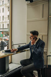 Businessman writing while talking on smart phone sitting at table in hotel lounge - MASF32157