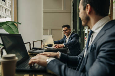 Glücklicher Geschäftsmann im Gespräch mit einem männlichen Kollegen, der einen Laptop benutzt, während er in einer Hotel-Lounge sitzt - MASF32158