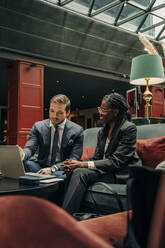Männlicher Berufstätiger, der mit einer Kollegin über einen Laptop diskutiert, während er in einer Hotellounge sitzt - MASF32183