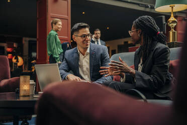 Smiling businessman looking at female colleague discussing while sitting in hotel lounge - MASF32188