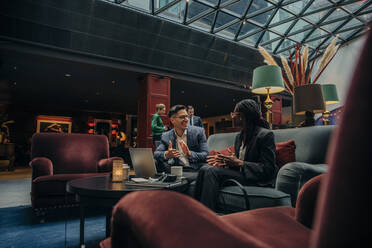 Smiling male professional discussing with female colleague sitting in hotel lounge - MASF32189